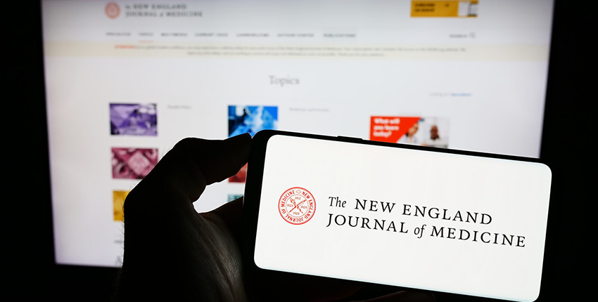 A hand holds a cellphone with the logo of The New England Journal of Medicine on screen, in front of a laptop screen displaying the journal's webpage — health tech coverage from STAT