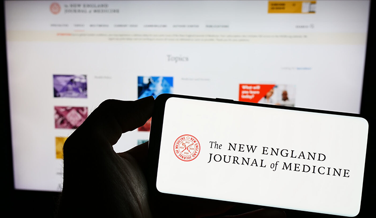 A hand holds a cellphone with the logo of The New England Journal of Medicine on screen, in front of a laptop screen displaying the journal's webpage — health tech coverage from STAT
