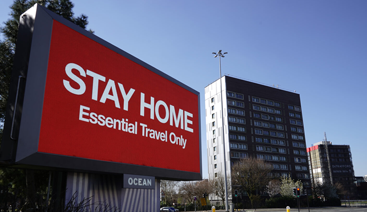 A digital board displays red background with white text that reads "STAY HOME" on the top and "Essential Travel Only" on the bottom — first opinion coverage from STAT